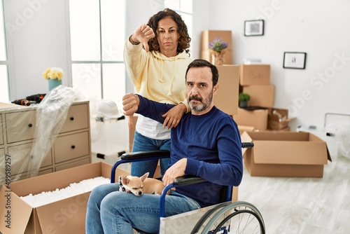 Middle age hispanic couple and dog sitting on wheelchair at new home with angry face, negative sign showing dislike with thumbs down, rejection concept