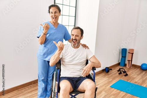 Hispanic middle age man sitting on wheelchair and nurse at rehabilitation clinic pointing thumb up to the side smiling happy with open mouth