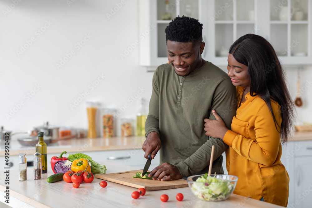 Young Black Woman Looking At Her Husband Cooking Salad In Kitchen Young Black Woman Looking At Her Husband Cooking Salad In Kitchen
