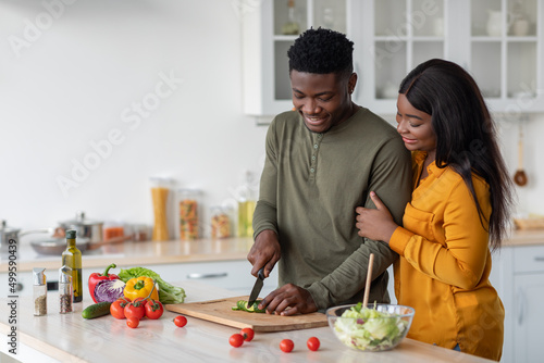 Young Black Woman Looking At Her Husband Cooking Salad In Kitchen