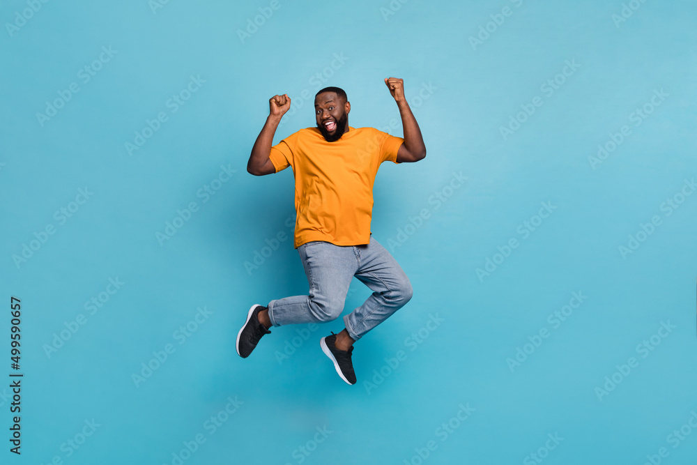 Photo of attractive lucky guy dressed orange t-shirt jumping high rising fists isolated blue color background