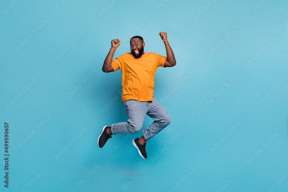 Photo of attractive lucky guy dressed orange t-shirt jumping high rising fists isolated blue color background Photo of attractive lucky guy dressed orange t-shirt jumping high rising fists isolated blue color background