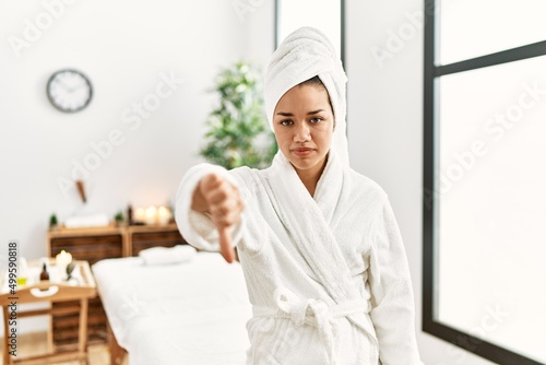 Young brunette woman wearing towel and bathrobe standing at beauty center looking unhappy and angry showing rejection and negative with thumbs down gesture. bad expression.