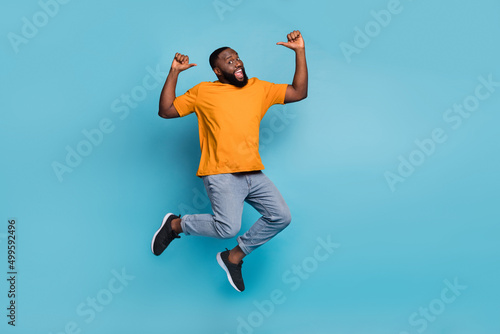 Photo of sweet excited guy wear orange t-shirt jumping high pointing thumbs himself isolated blue color background