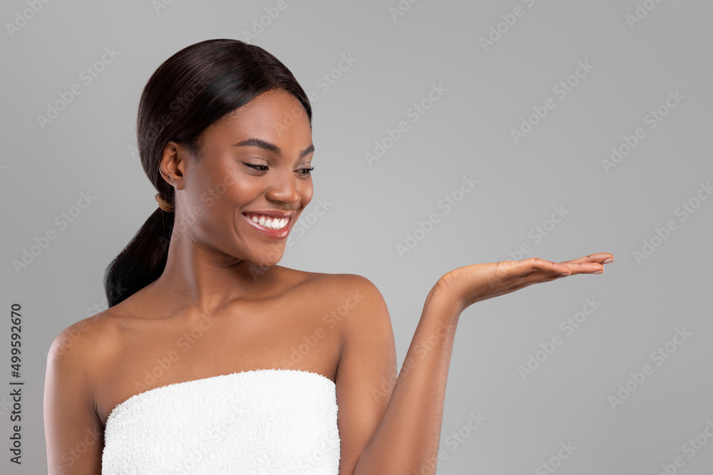 Smiling beautiful black lady in towel looking at hand with blank space, isolated on gray background Smiling beautiful black lady in towel looking at hand with blank space, isolated on gray background