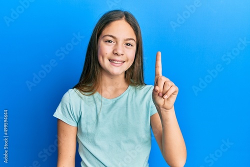Beautiful brunette little girl wearing casual white t shirt showing and pointing up with finger number one while smiling confident and happy.