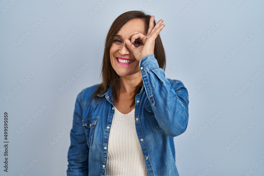 Middle age brunette woman wearing casual denim jacket over isolated background doing ok gesture with hand smiling, eye looking through fingers with happy face. Middle age brunette woman wearing casual denim jacket over isolated background doing ok gesture with hand smiling, eye looking through fingers with happy face.