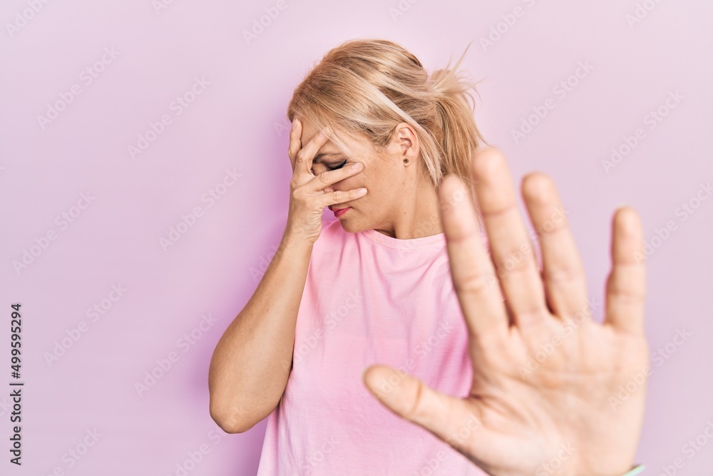 Young blonde woman wearing casual pink t shirt covering eyes with hands and doing stop gesture with sad and fear expression. embarrassed and negative concept. Young blonde woman wearing casual pink t shirt covering eyes with hands and doing stop gesture with sad and fear expression. embarrassed and negative concept.