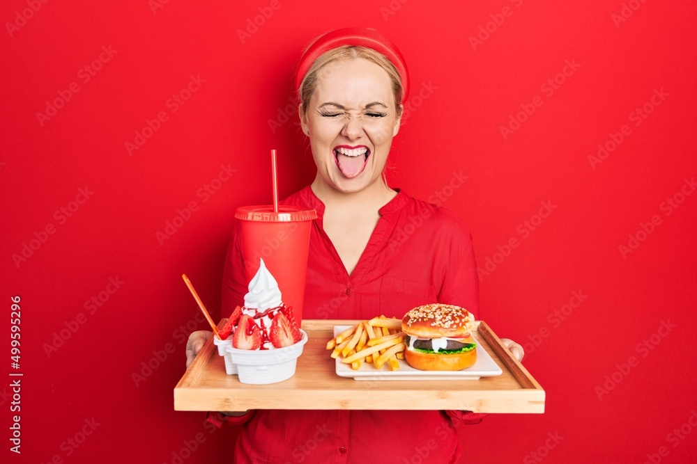 Young blonde woman eating a tasty classic burger with fries and soda sticking tongue out happy with funny expression. Young blonde woman eating a tasty classic burger with fries and soda sticking tongue out happy with funny expression.