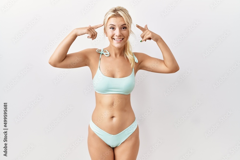 Young caucasian woman wearing bikini over isolated background smiling pointing to head with both hands finger, great idea or thought, good memory Young caucasian woman wearing bikini over isolated background smiling pointing to head with both hands finger, great idea or thought, good memory