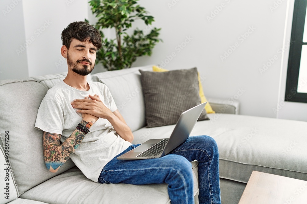 Hispanic man with beard sitting on the sofa smiling with hands on chest with closed eyes and grateful gesture on face. health concept.