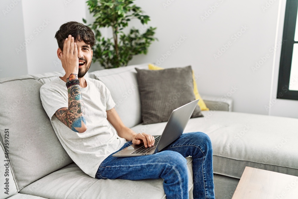 Hispanic man with beard sitting on the sofa covering one eye with hand, confident smile on face and surprise emotion.
