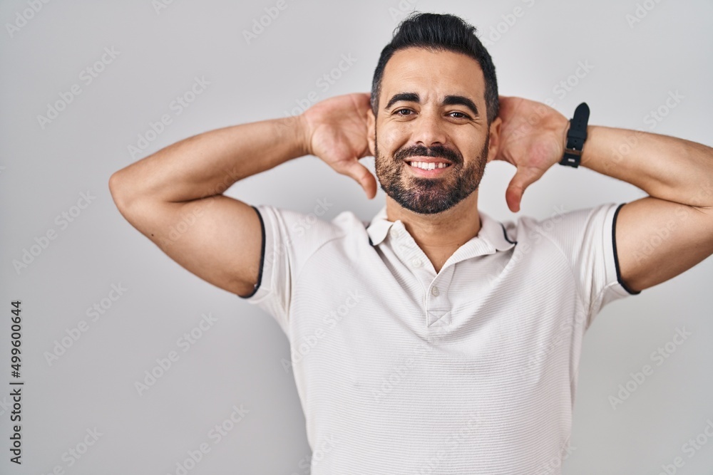 Young hispanic man with beard wearing casual clothes over white background relaxing and stretching, arms and hands behind head and neck smiling happy Young hispanic man with beard wearing casual clothes over white background relaxing and stretching, arms and hands behind head and neck smiling happy