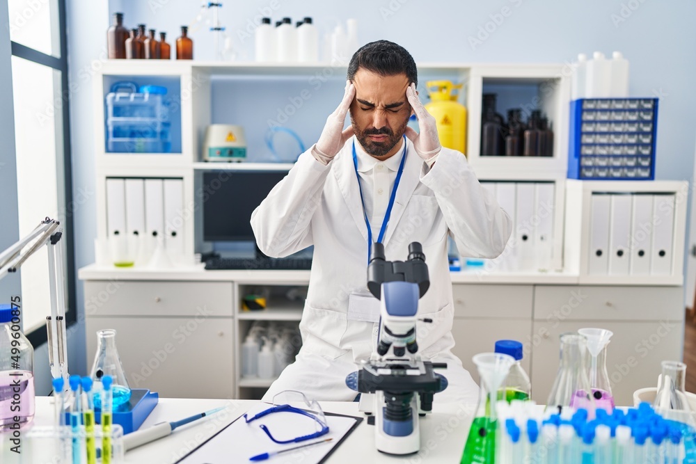 Young hispanic man with beard working at scientist laboratory with hand on head for pain in head because stress. suffering migraine. Young hispanic man with beard working at scientist laboratory with hand on head for pain in head because stress. suffering migraine.