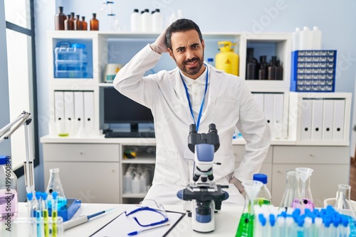 Young hispanic man with beard working at scientist laboratory confuse and wonder about question. uncertain with doubt, thinking with hand on head. pensive concept.