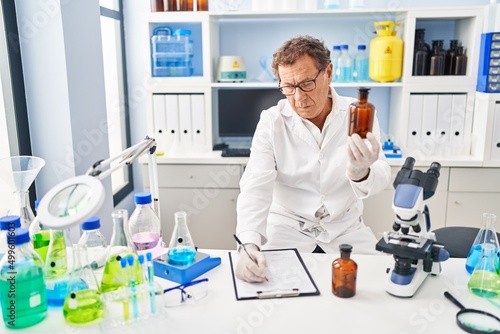 Middle age man wearing scientist uniform holding bottle writing on clipboard at laboratory