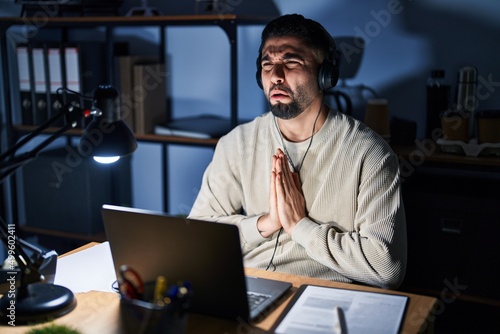 Foto Young handsome man working using computer laptop at night begging and praying with hands together with hope expression on face very emotional and worried