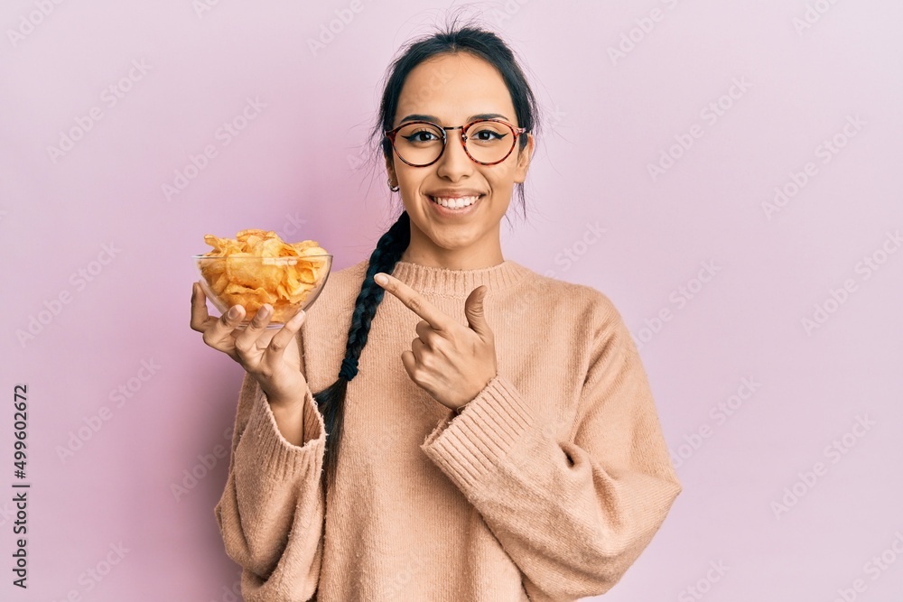 Young hispanic girl holding potato chip smiling happy pointing with hand and finger Young hispanic girl holding potato chip smiling happy pointing with hand and finger