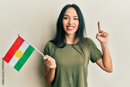 Young hispanic girl holding kurdistan flag smiling with an idea or question pointing finger with happy face, number one