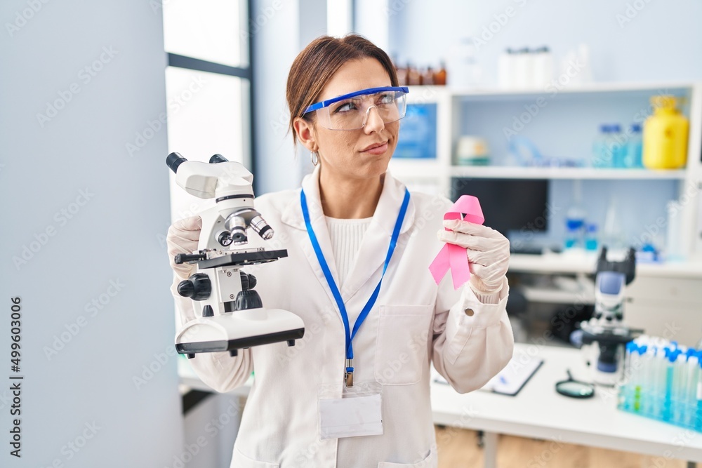 Young brunette woman working at scientist laboratory holding pink ribbon smiling looking to the side and staring away thinking.