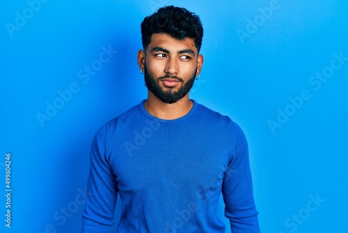 Arab man with beard wearing casual blue sweater smiling looking to the side and staring away thinking.