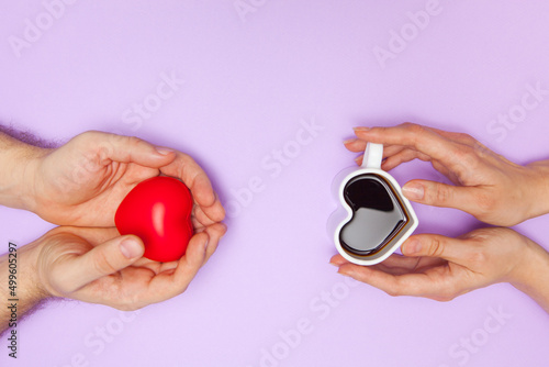 Woman's hands holds heart shaped cup of coffee and man's hands holds red heart. Black hot coffee in a white heart-shaped cup.