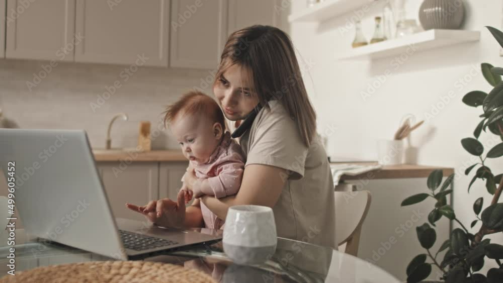 Medium of brown-haired young Biracial woman holding cute baby daughter, talking on phone, sitting at table, using laptop computer in kitchen in afternoon Medium of brown-haired young Biracial woman holding cute baby daughter, talking on phone, sitting at table, using laptop computer in kitchen in afternoon