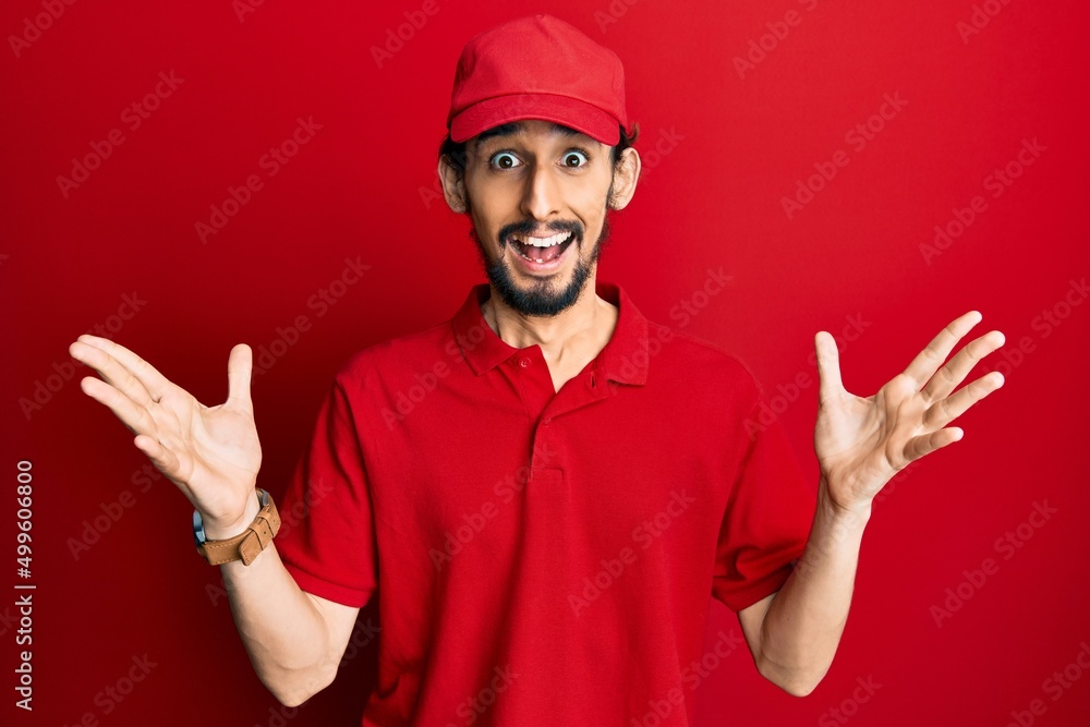 Young hispanic man wearing delivery uniform and cap celebrating victory with happy smile and winner expression with raised hands Young hispanic man wearing delivery uniform and cap celebrating victory with happy smile and winner expression with raised hands