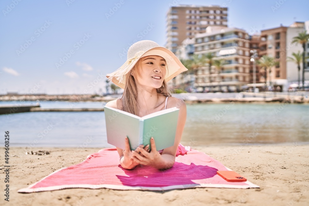 Young chinese girl reading book lying on the towel at the beach.