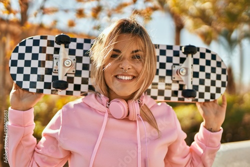 Young blonde skater girl smiling happy holding skate at the city.