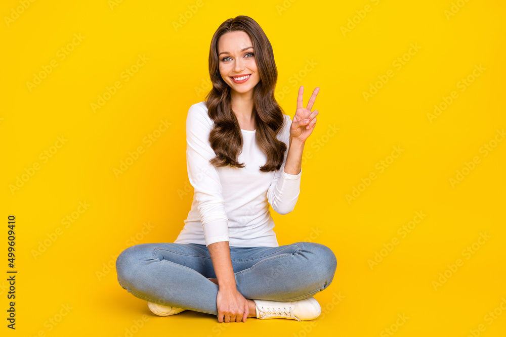 Photo of peaceful adorable woman wear white shirt showing v-sign sitting legs crossed isolated yellow color background Photo of peaceful adorable woman wear white shirt showing v-sign sitting legs crossed isolated yellow color background