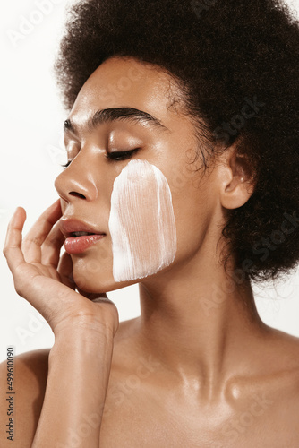 Vertical portrait of sensual black woman, gently touching her soft, healthy face with facial cream, skincare product on cheek, using lotion or daily care for better skin tone, pink background
