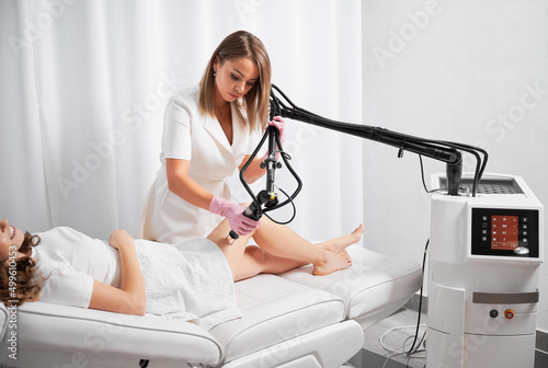 Dermatologist working with modern laser device for polishing and repairing skin defects. Beautician demonstrating work of CO2 laser, warming skin of clients thigh, stimulating local rejuvenation. photo