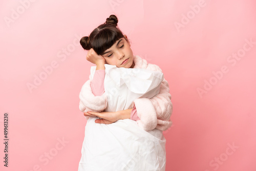 Little caucasian girl isolated on pink background in pajamas and holding a pillow while sleeping