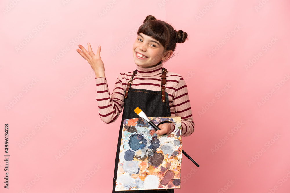 Little artist girl holding a palette isolated on pink background extending hands to the side for inviting to come Little artist girl holding a palette isolated on pink background extending hands to the side for inviting to come