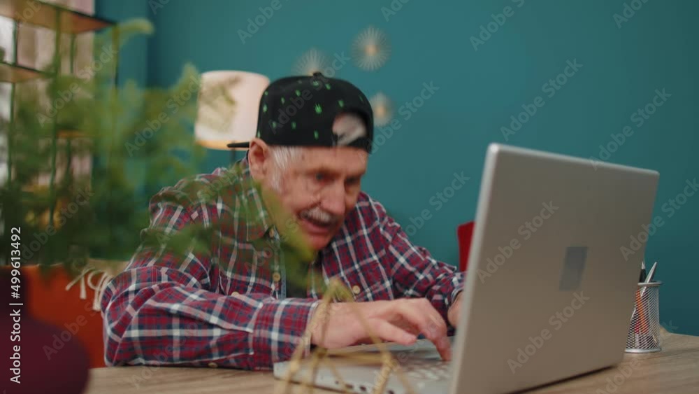 Elderly old grandfather man in stylish cap playing computer video games on laptop computer, wins, rises hands, celebrate, smiles happily. Gamer, having fun, rest. Virtual reality. Modern technologies Elderly old grandfather man in stylish cap playing computer video games on laptop computer, wins, rises hands, celebrate, smiles happily. Gamer, having fun, rest. Virtual reality. Modern technologies