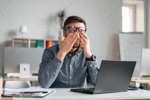 Mature male employee rubbing tired irritated eyes, working with laptop too much, exhausted from online job at office