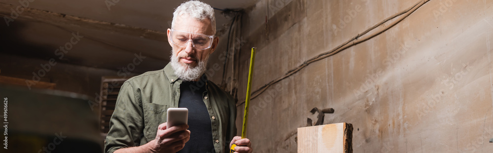 bearded carpenter with tape measure using smartphone in workshop, banner. bearded carpenter with tape measure using smartphone in workshop, banner.