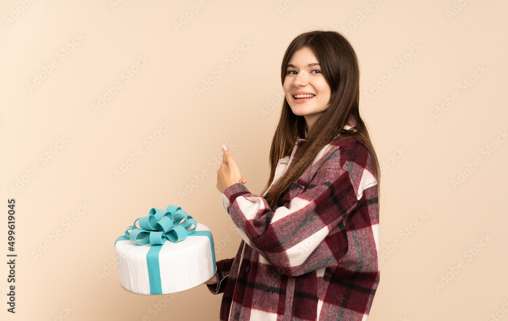 Young Ukrainian girl holding a big cake isolated on beige background pointing back Young Ukrainian girl holding a big cake isolated on beige background pointing back