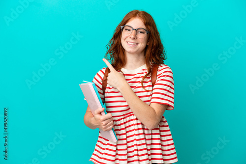 Student teenager redhead girl isolated on blue background pointing to the side to present a product