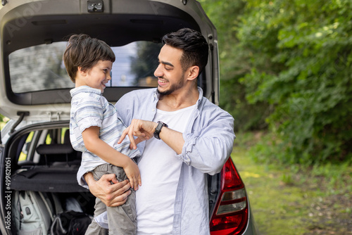 Happy arab man holding son in arms and playing with him near car with open trunk outdoor in summer #499621051