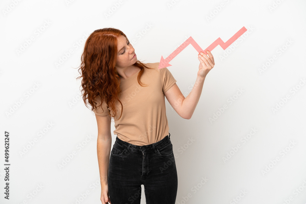 Teenager redhead girl isolated on white background holding a downward arrow and with sad expression Teenager redhead girl isolated on white background holding a downward arrow and with sad expression