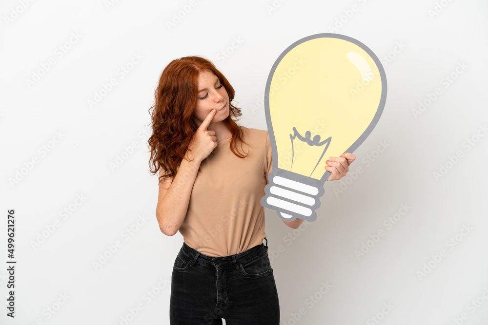 Teenager redhead girl isolated on white background holding a bulb icon and having doubts Teenager redhead girl isolated on white background holding a bulb icon and having doubts