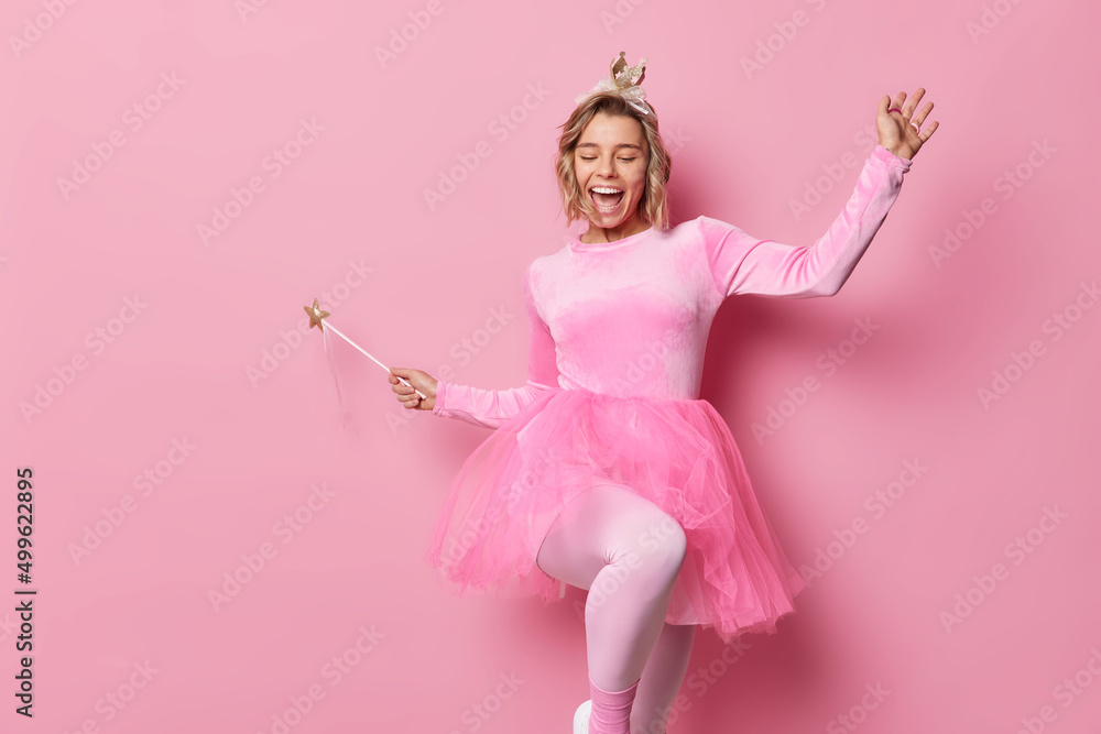 Cheerful young woman dances carefree has upbeat mood wears small crown and dress holds magic wand entertains children on party moves against pink background blank copy space for your promotion Cheerful young woman dances carefree has upbeat mood wears small crown and dress holds magic wand entertains children on party moves against pink background blank copy space for your promotion