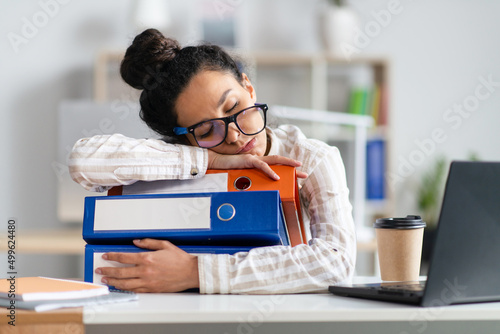 Tired businesswoman sleeping on stack of folders at her desk in front of laptop, feeling exhausted at office photo