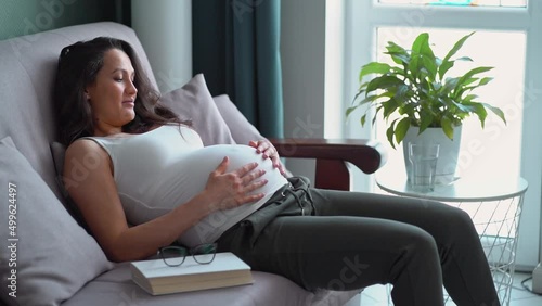 Pregnant middle age woman sit on sofa in modern living room looking and caressing her belly with love Pregnancy, maternity concept. Copy space. Slow motion