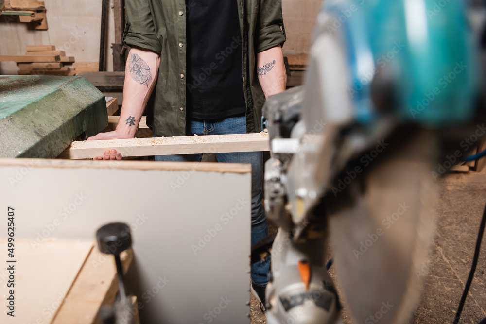 partial view of tattooed woodworker holding board while working on blurred foreground.