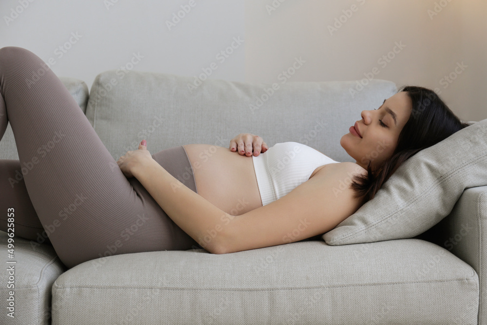 Young beautiful woman on second trimester of pregnancy. Close up of pregnant female on the couch in yoga pants with arms on her round belly. Expecting a child concept. Background, copy space. Young beautiful woman on second trimester of pregnancy. Close up of pregnant female on the couch in yoga pants with arms on her round belly. Expecting a child concept. Background, copy space.