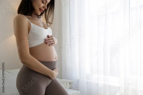 Young beautiful woman on second trimester of pregnancy. Close up of pregnant female in yoga pants with arms on her round belly. Expecting a child concept. Background, copy space.