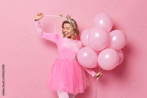 Photo of glad young European woman has fun on party poses with bunch of helium balloons uses magic wand to make your dreams come true poses against pink background. People and holidays concept